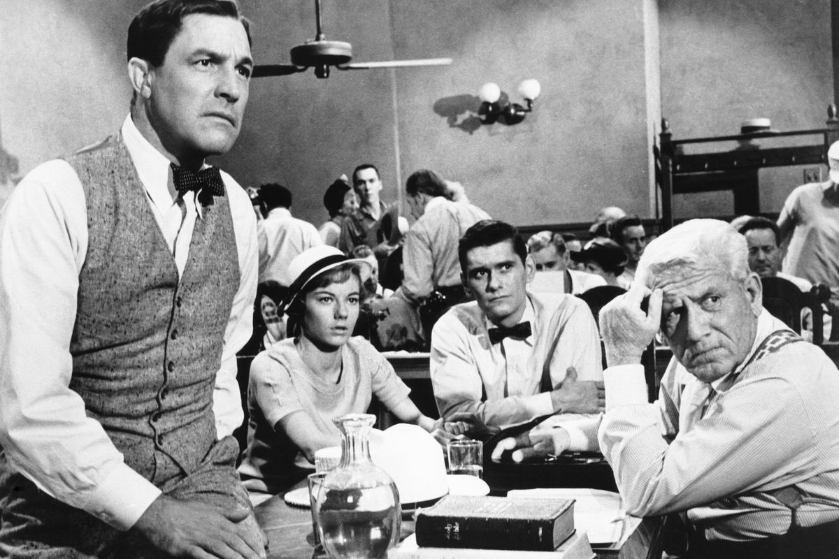 LtR: Gene Kelly as E. K. Hornbeck, Donna Anderson as Rachel Brown, Dick York as Bertram T. Cates, and Spencer Tracy as Henry Drummond in Inherit the Wind. They’re all looking concerned as they sit around the defendant’s table in a packed courtroom (Hornbeck is leaning with one leg up on the table. Brown has a cute little sunhat on and the men are in their shirtsleeves despite the formal setting.