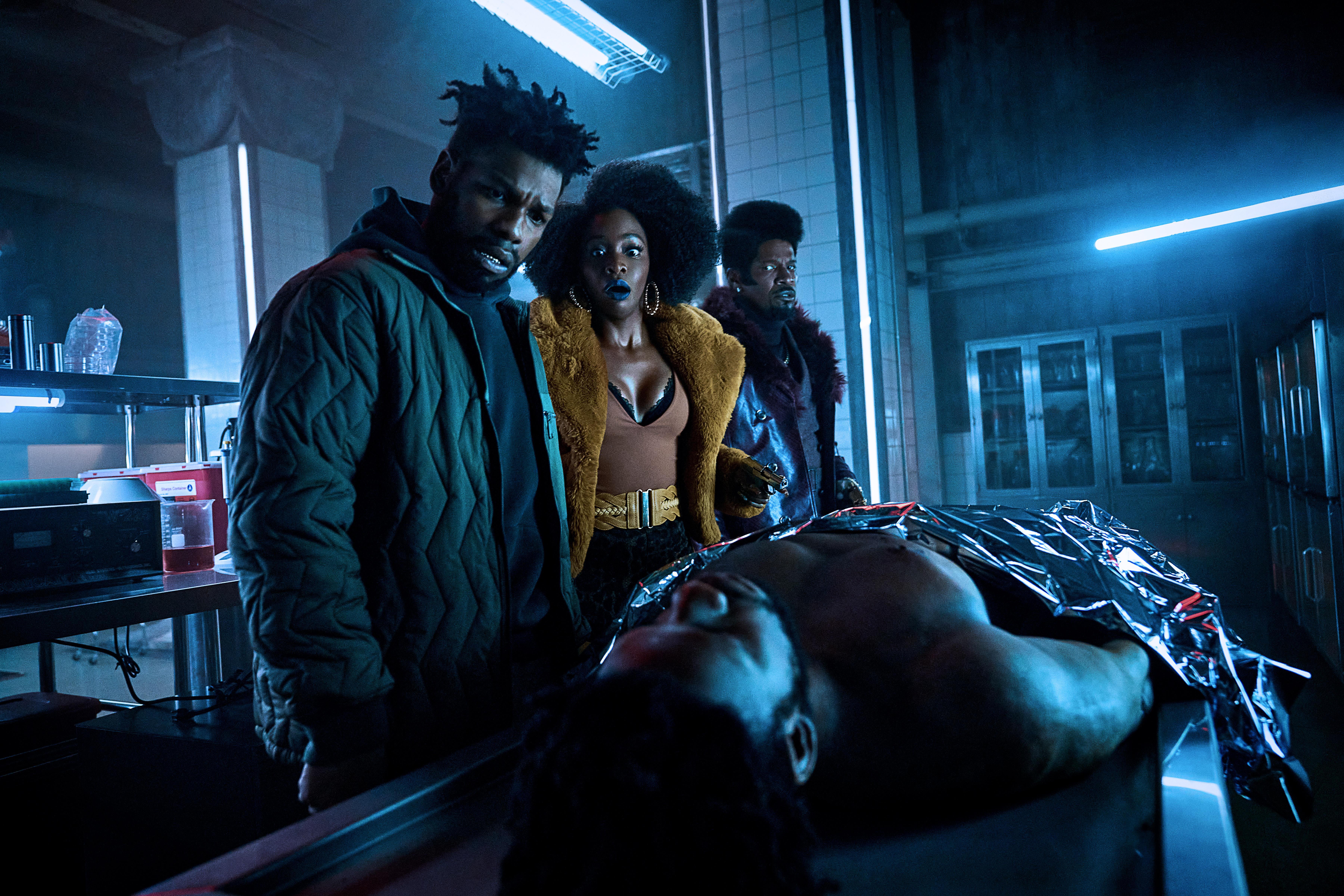 (L to R) John Boyega as Fontaine, Teyonah Parris as Yo-Yo and Jamie Foxx as Slick Charles staring down at a dead body on a examination table in They Cloned Tyrone.